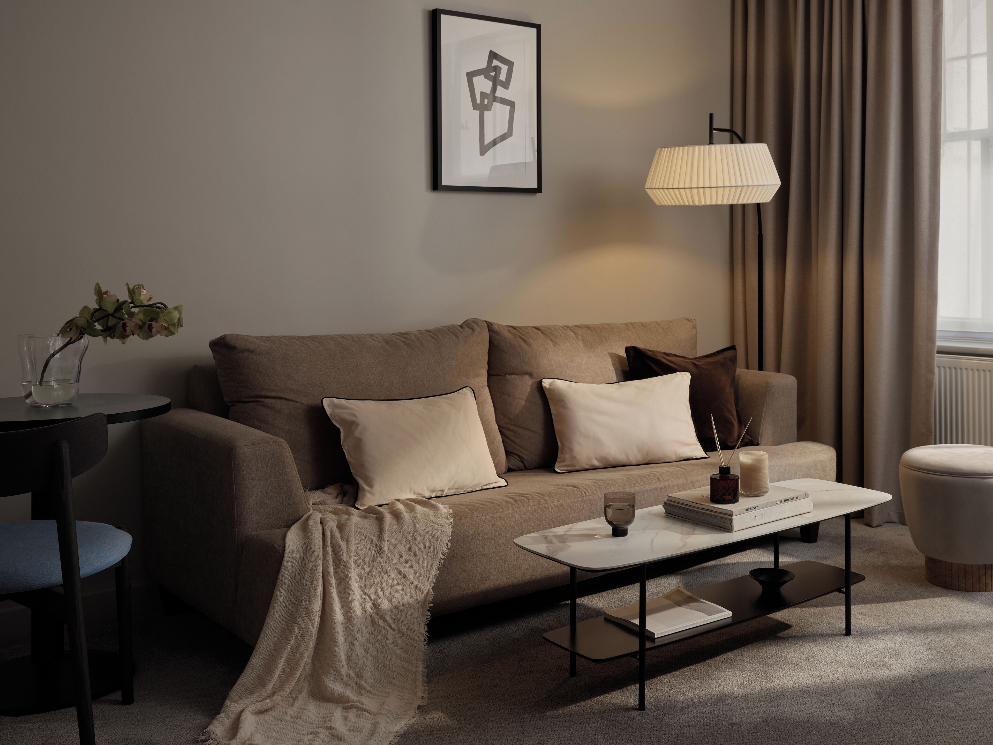Ein modernes Wohnzimmer mit einem beigen Sofa, einem Couchtisch, einem Beistelltisch mit Vase und Kerzen sowie einer Lampe.