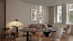 Un salon moderne avec une table basse ronde, un canapé et plusieurs fauteuils. Deux œuvres d’art encadrées ornent le mur, ainsi qu’une lampe sur pied et une fenêtre donnant sur un paysage verdoyant.