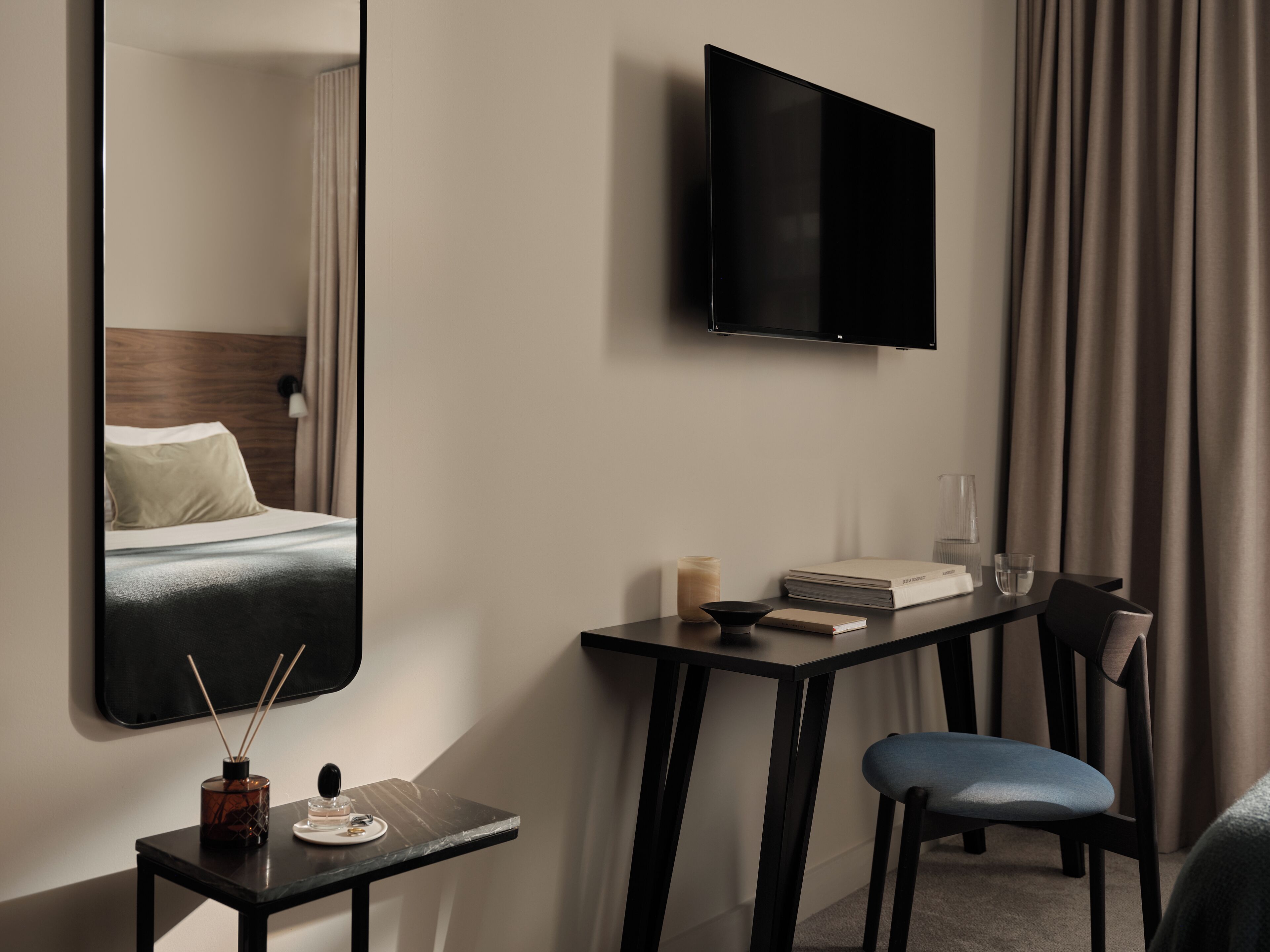 Una habitación de hotel moderna con una cama, un televisor montado en la pared, una mesita de noche con una lámpara, una silla y un escritorio con su respectiva silla.