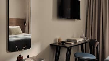 Una habitación de hotel moderna con una cama, un televisor montado en la pared, una mesita de noche con una lámpara, una silla y un escritorio con su respectiva silla.