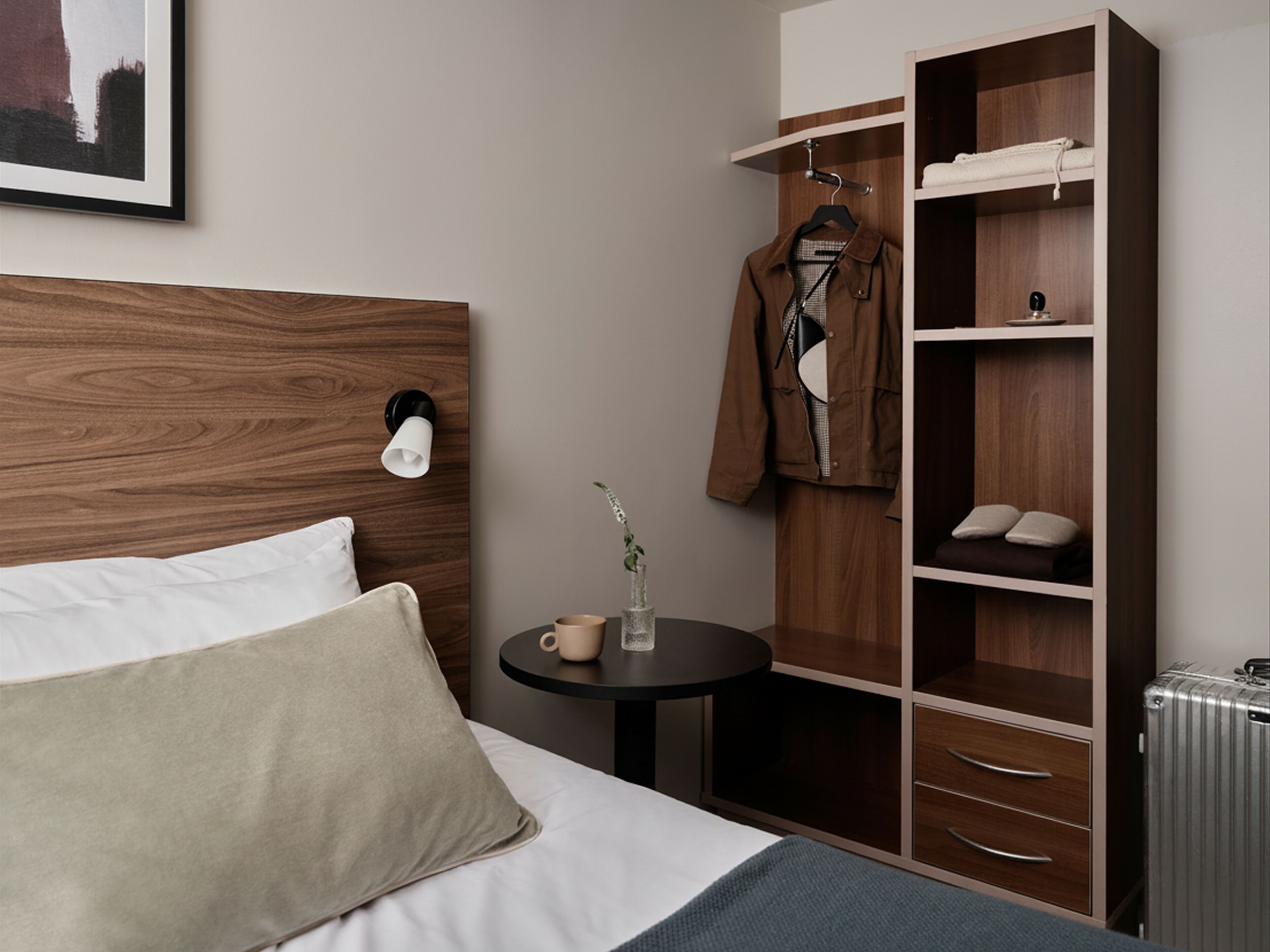 Een keurig ingerichte hotelkamer met een houten hoofdeinde, een bed met kussens, een klein rond tafeltje met een kop en vaas, een kledingkast met hangende kleding en een radiator.