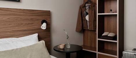 Een keurig ingerichte hotelkamer met een houten hoofdeinde, een bed met kussens, een klein rond tafeltje met een kop en vaas, een kledingkast met hangende kleding en een radiator.