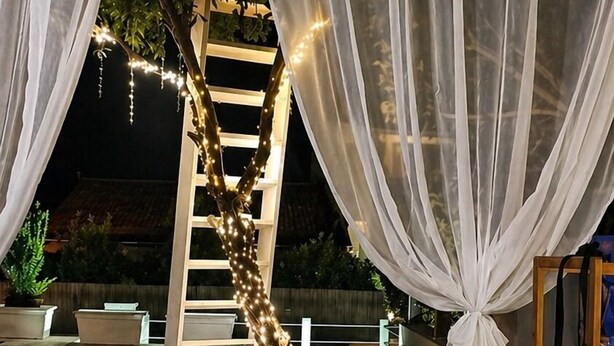 Um pátio externo com deck de madeira, cortinas brancas, escada de madeira e luzes de fada.