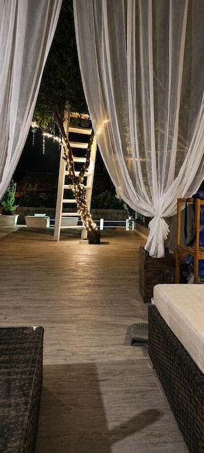 An outdoor patio with a wooden deck, white drapes, a wooden ladder, and string lights.