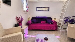 Quarto com sofá roxo, cadeiras de vime, uma mesinha com telefone e um espelho decorativo.