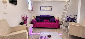 Une pièce avec un canapé violet, des fauteuils en osier, une petite table avec un téléphone et un miroir décoratif.