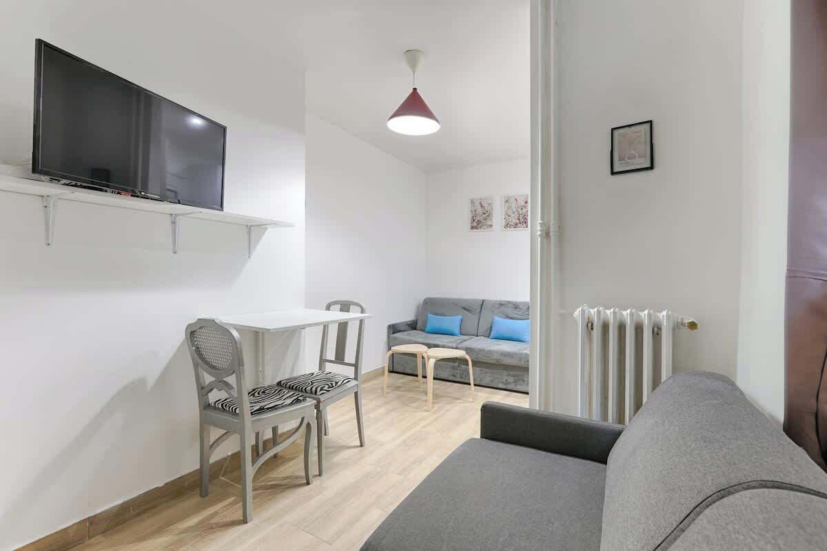 A compact living space with a flat-screen TV mounted on the wall, a dining area with a table and chairs, a grey sofa, and a radiator.