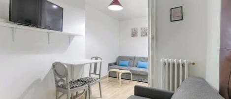 A compact living space with a flat-screen TV mounted on the wall, a dining area with a table and chairs, a grey sofa, and a radiator.
