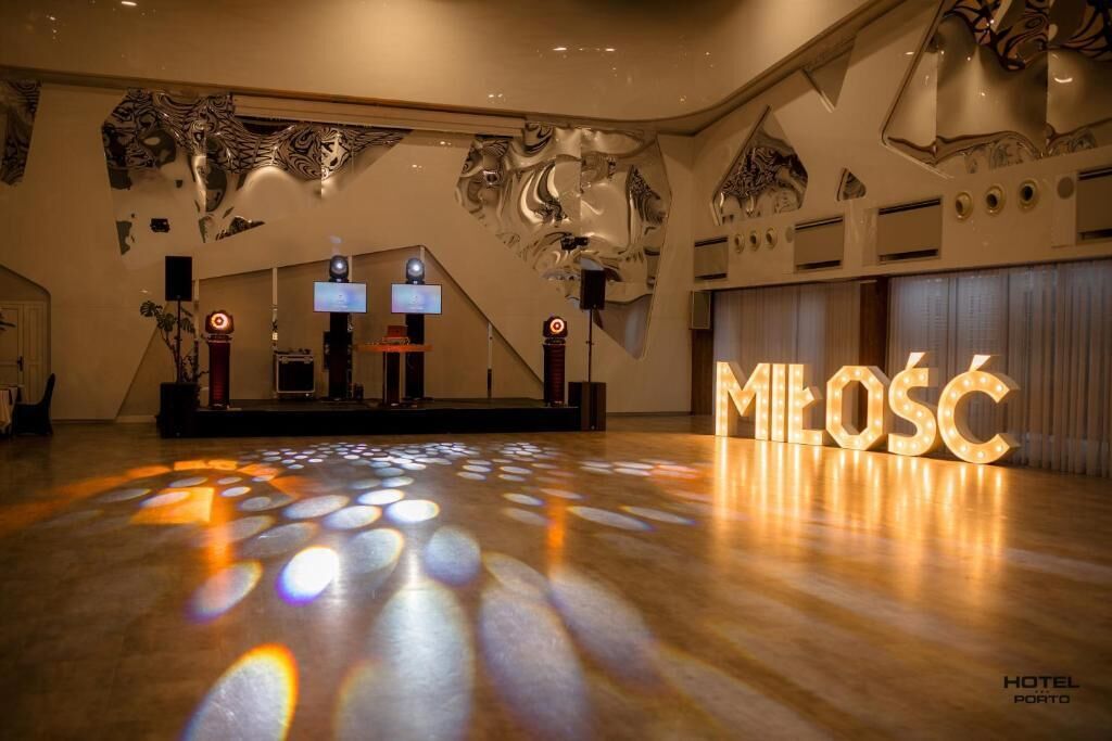 Un hall d’entrée spacieux avec un plancher en bois, une scène équipée de haut-parleurs et un grand panneau lumineux portant l’inscription « MILOSC ».