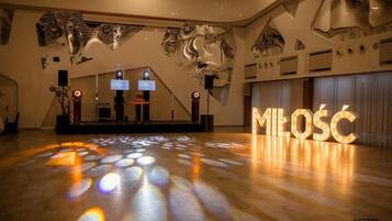 Un hall d’entrée spacieux avec un plancher en bois, une scène équipée de haut-parleurs et un grand panneau lumineux portant l’inscription « MILOSC ».