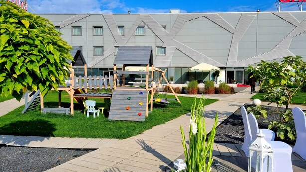 A playground with climbing structures and a seating area, surrounded by a well-maintained lawn and a building with a geometric facade. - HOTEL PORTO (Radomsko)