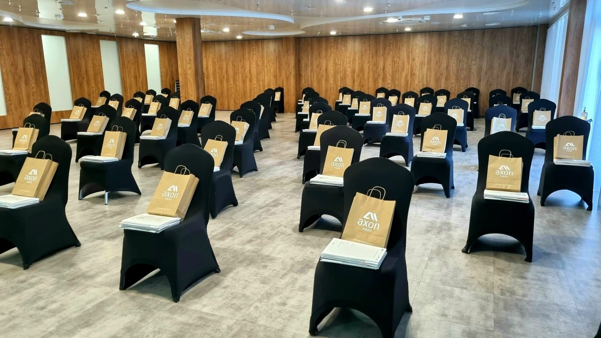 Une salle de conférence avec des rangées de chaises noires, chacune contenant un sac en papier brun étiqueté « axon media ».