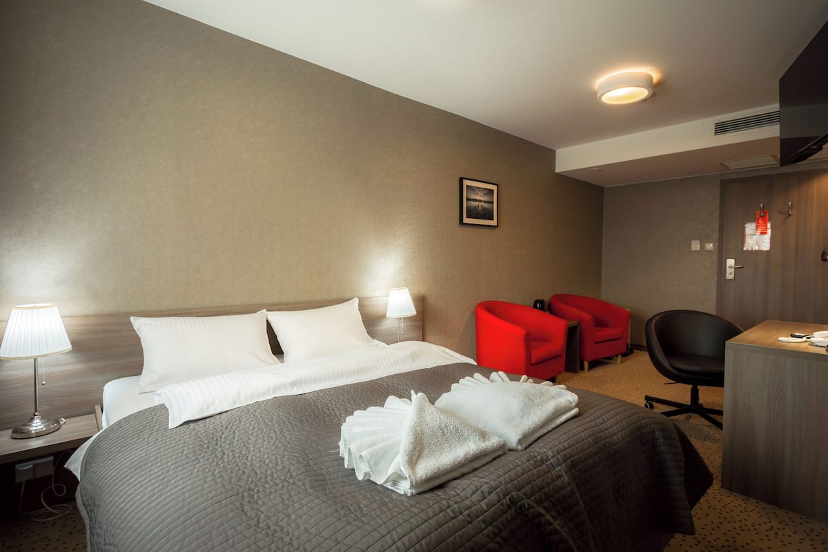 A hotel room with a bed, two bedside lamps, a desk with a chair, and two red armchairs.