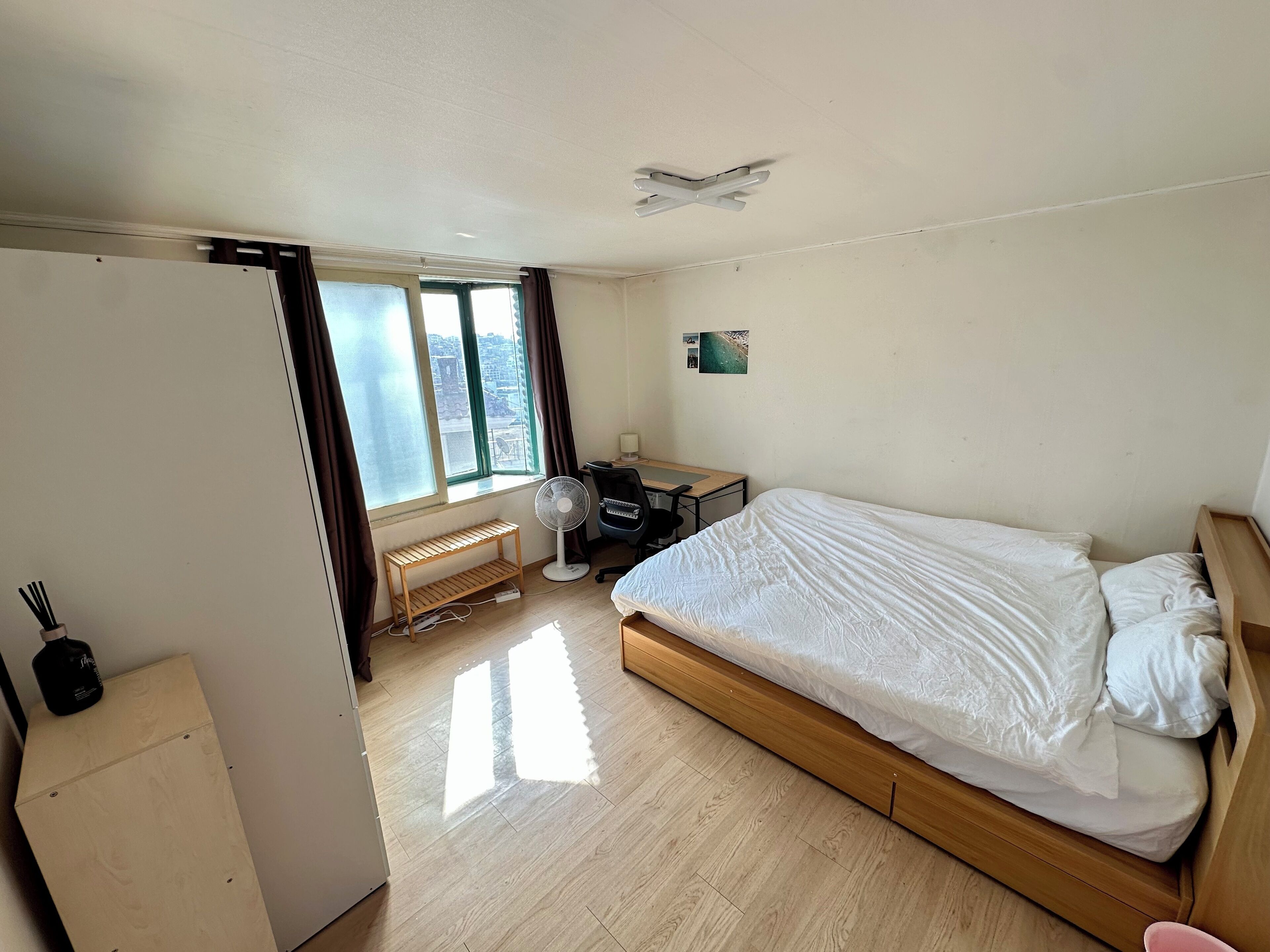 Een slaapkamer met een bed, een bureau, een stoel, een ventilator en een raam met gordijnen.