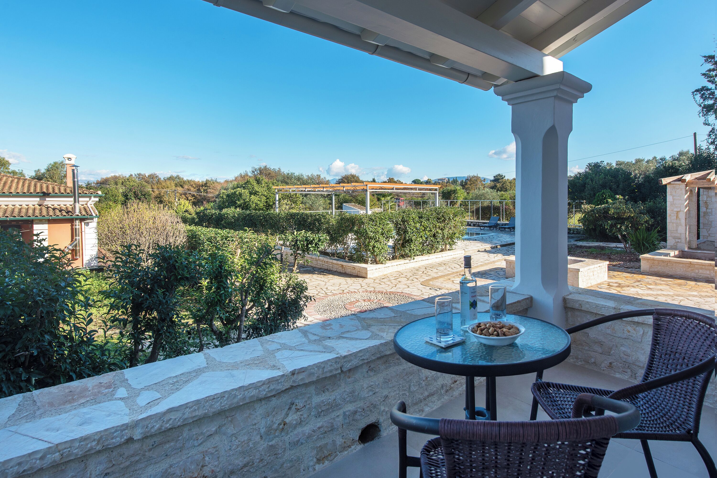 A balcony with a stone wall, a round table with chairs, and a view of a garden and buildings in the background.
