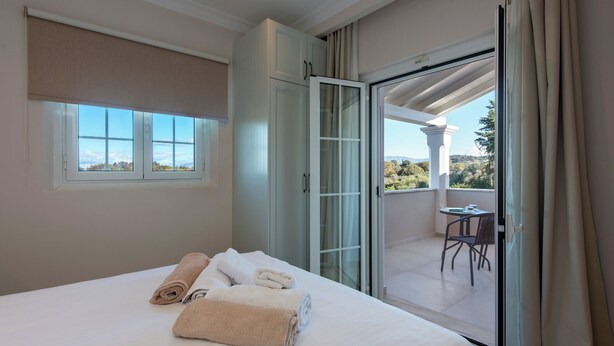 A hotel room with a bed, a large window, a sliding glass door leading to a balcony, and a view of the outdoors. - Nancy Suites (Kassiopi)