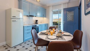 A compact kitchen with blue cabinets, a white refrigerator, and a dining table set for breakfast. - Nancy Suites (Kassiopi)