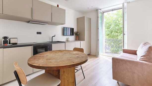 A modern kitchen with a round wooden table, white cabinets, and a large window offering a view of greenery. - Charmant Studio II au coeur de Vincennes (Vincennes)