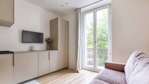 A modern living room with a sofa, a flat-screen TV mounted on the wall, and a sliding glass door leading to a balcony. - Charmant Studio II au coeur de Vincennes (Vincennes)