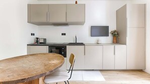 A modern kitchen with a wooden dining table, white cabinets, a built-in oven, and a flat-screen TV mounted above the counter. - Charmant Studio II au coeur de Vincennes (Vincennes)