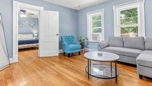 Un salón con piso de madera, un sofá gris, un sillón azul, una mesa de centro redonda de vidrio y un ventilador de techo.
