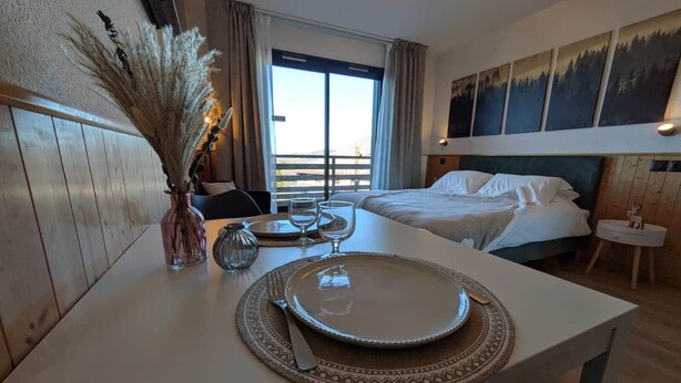 A hotel room with a dining area set for two, a bed with white linens, a view of the outdoors through a large window, and a painting on the wall. - Juracîmes Duo apartment for 2 people (Mijoux)