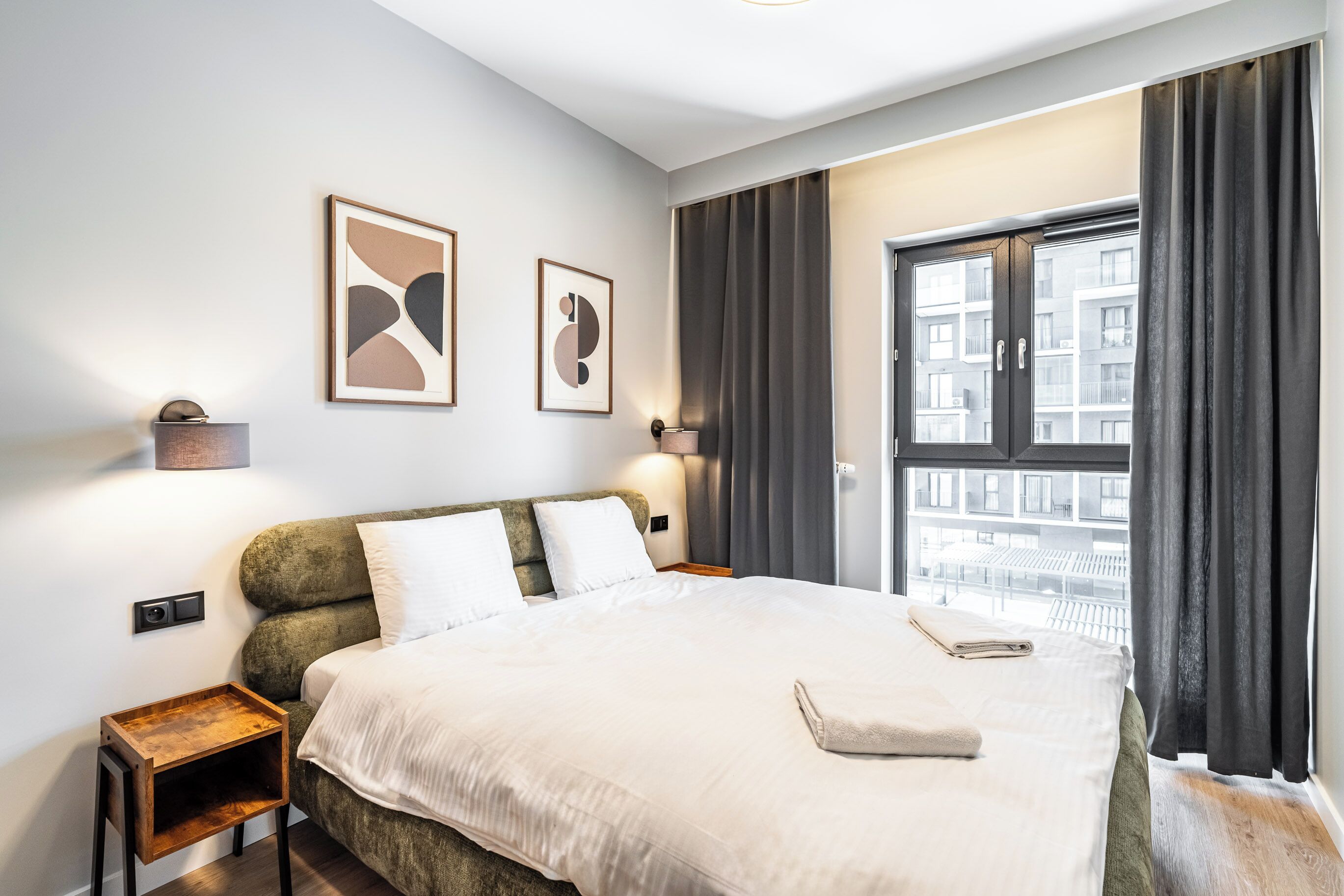 Moderni hotellihuone, jossa on suuri sänky, kaksi yöpöytää, ikkuna verhoilla ja abstraktia taidetta seinällä.
