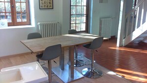 A dining area with a wooden table and four chairs, a radiator, and a window with a view of greenery. - Villa La Ballière: Where memories are written between heaven and earth (Coullons)