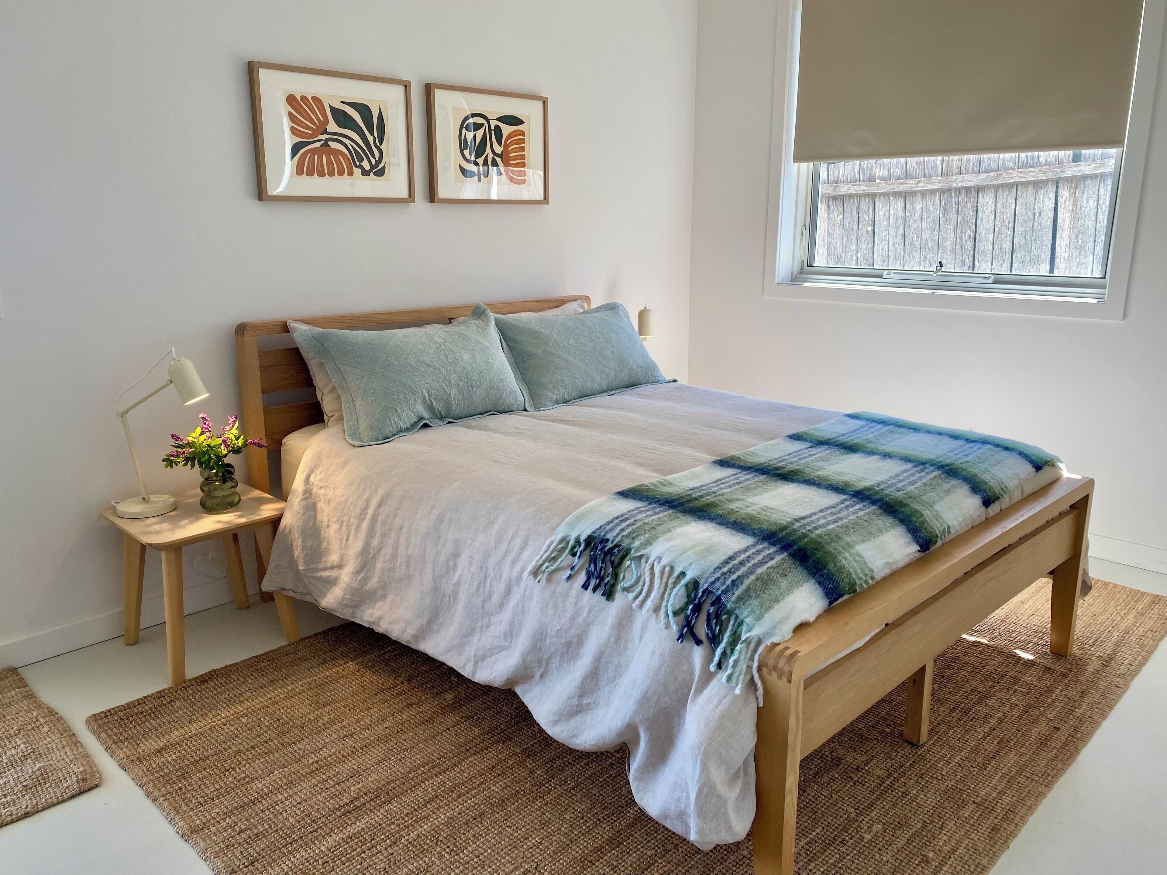 A bedroom with a wooden bed, a nightstand with a lamp and vase, two framed artworks on the wall, and a window with a view.