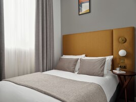 A neatly made bed with a headboard, a bedside table with a lamp, and a window with curtains.
