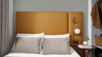 A neatly made bed with two pillows, a bedside table with a lamp, and a coat rack with a jacket.