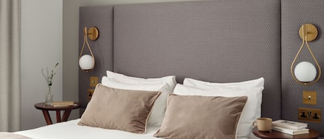 Una cama bien hecha con almohadas blancas y marrones, una manta color beige y una mesita de noche con un libro y una taza.