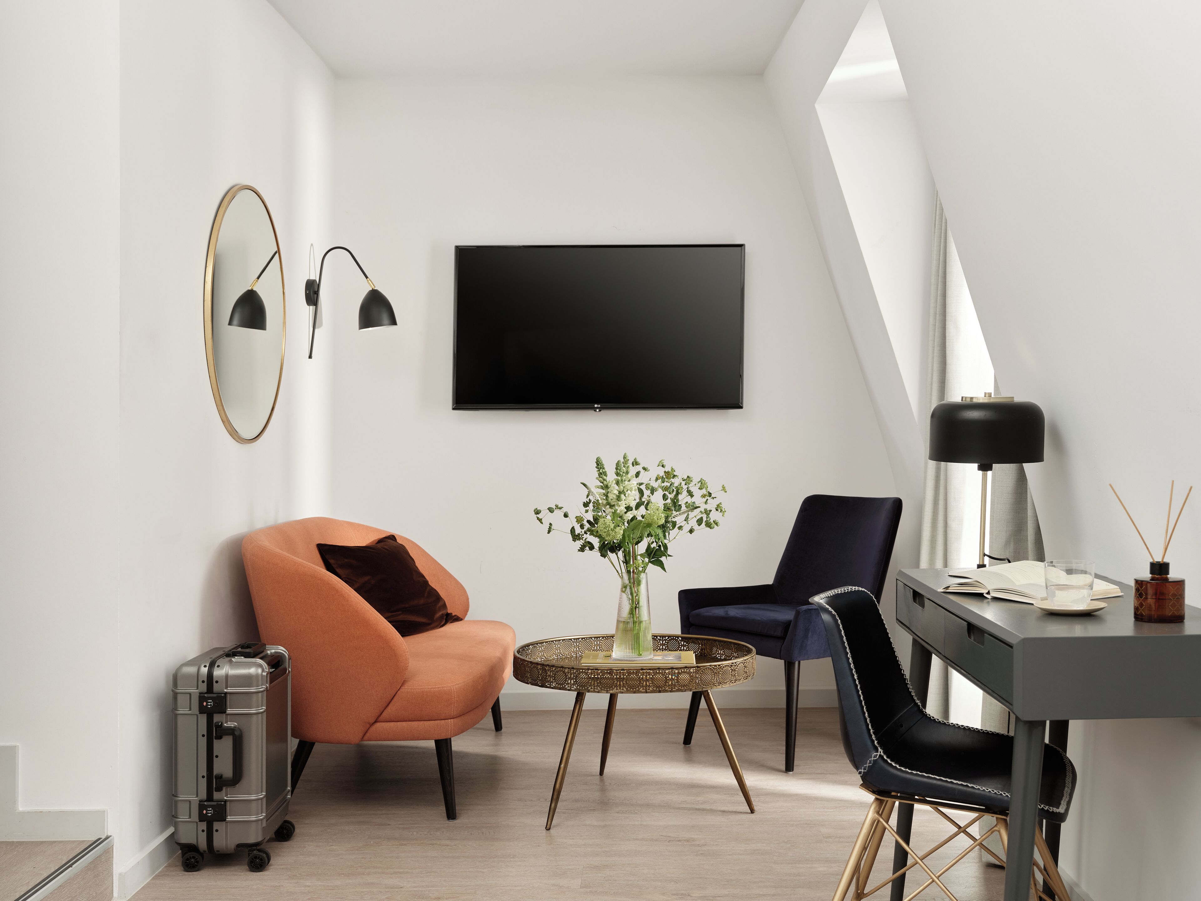 A modern living room with a flat-screen TV, two chairs, a small table with a vase, and a suitcase.