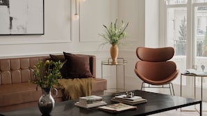 Una sala de estar moderna con un sofá de cuero, una mesa de centro con libros, un jarrón con flores y vistas a un paisaje urbano a través de grandes ventanales.