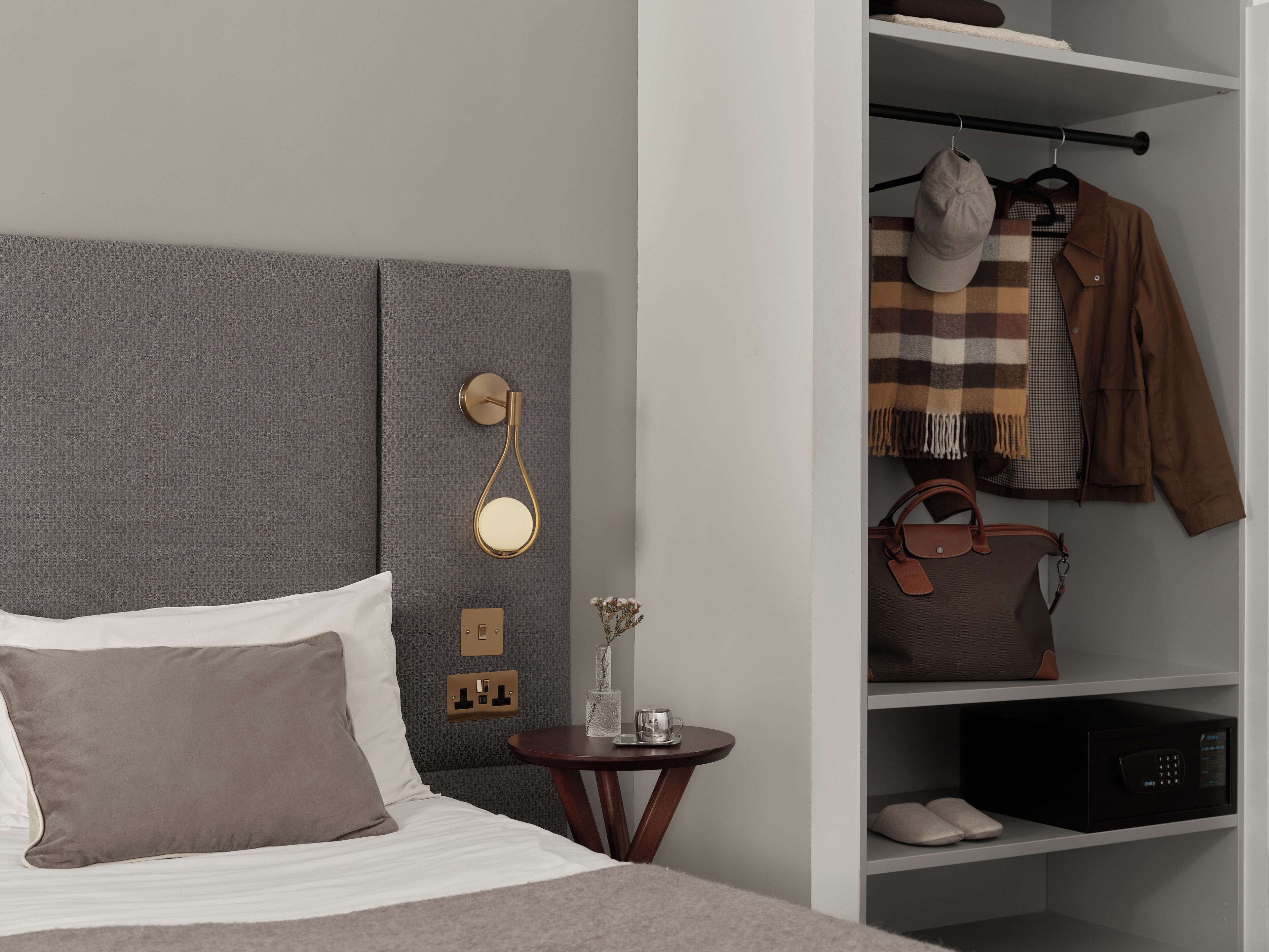 Een netjes ingerichte slaapkamer met een grijs hoofdeinde, een nachtkastje met een vaas en een kopje, en een garderobe met hangende kleding en schoenen.