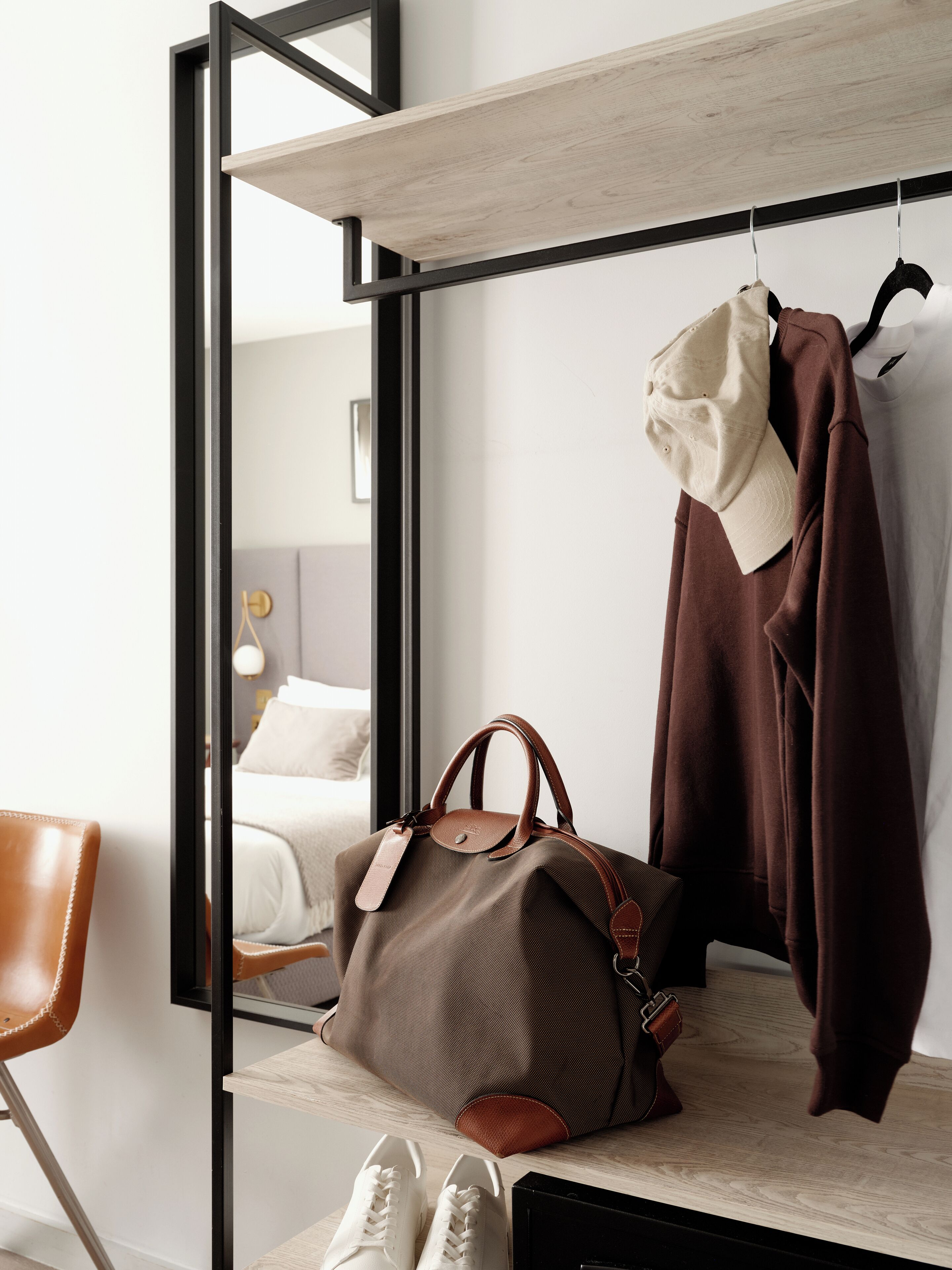 Una camera d'albergo con un letto, una mensola in legno, un appendiabiti e una borsa marrone, oltre a un paio di scarpe da ginnastica bianche.