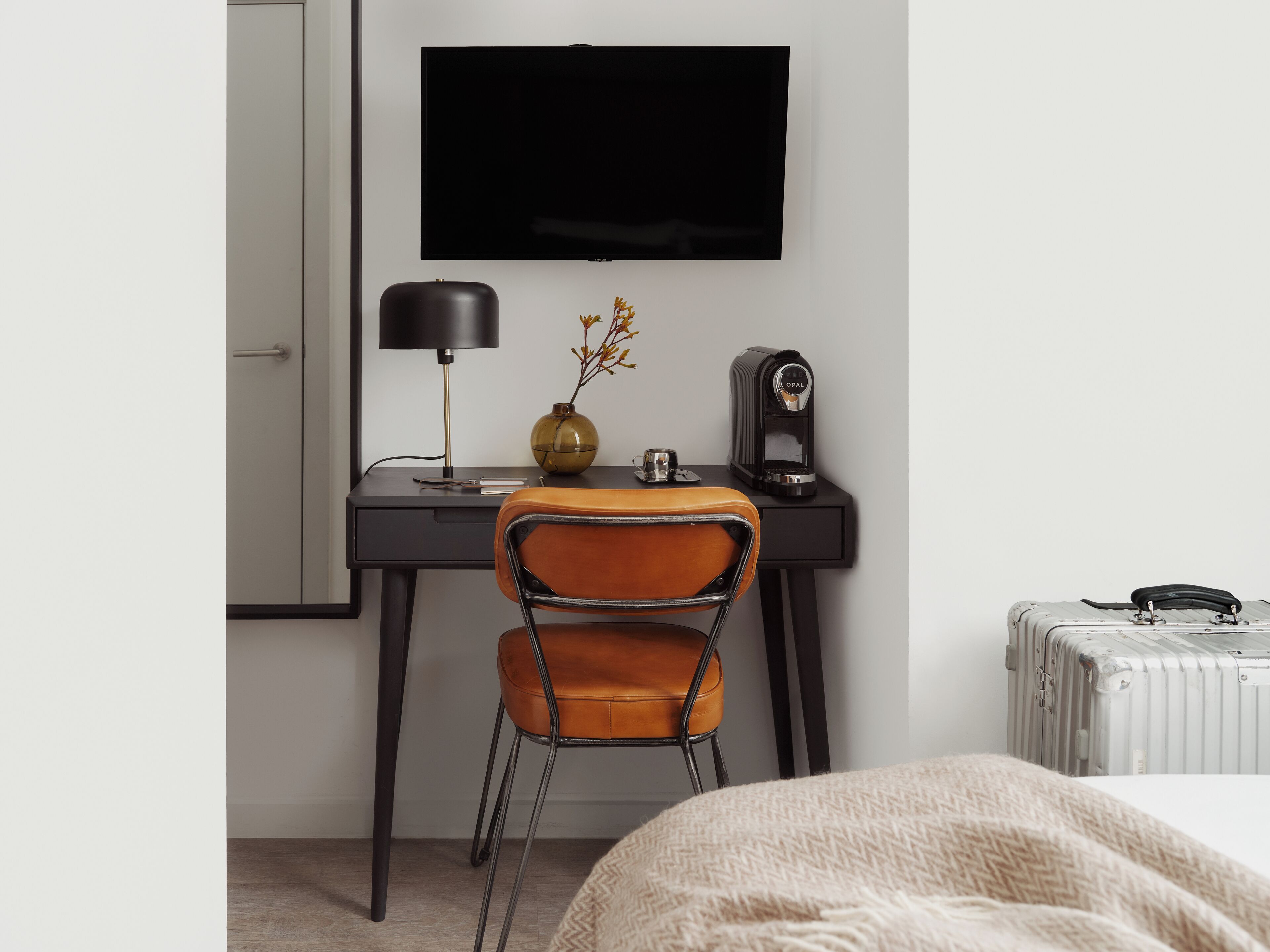 Ett hotellrum med en säng, ett skrivbord med en bärbar dator, en kaffebryggare, en stol och en resväska.