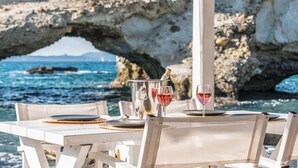 A table set for two with wine glasses and a bottle, overlooking a rocky coastline with a natural arch formation. - Aqua Houses (Milos)