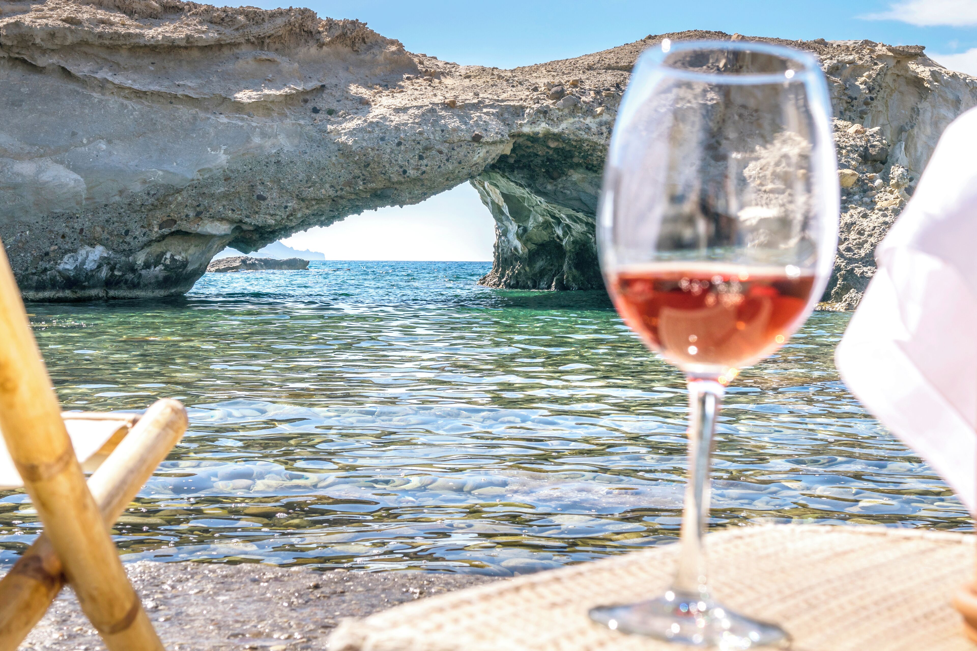 Una copa de vino tinto en una playa con agua cristalina y una formación rocosa al fondo.