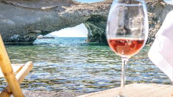 Una copa de vino tinto en una playa con agua cristalina y una formación rocosa al fondo.