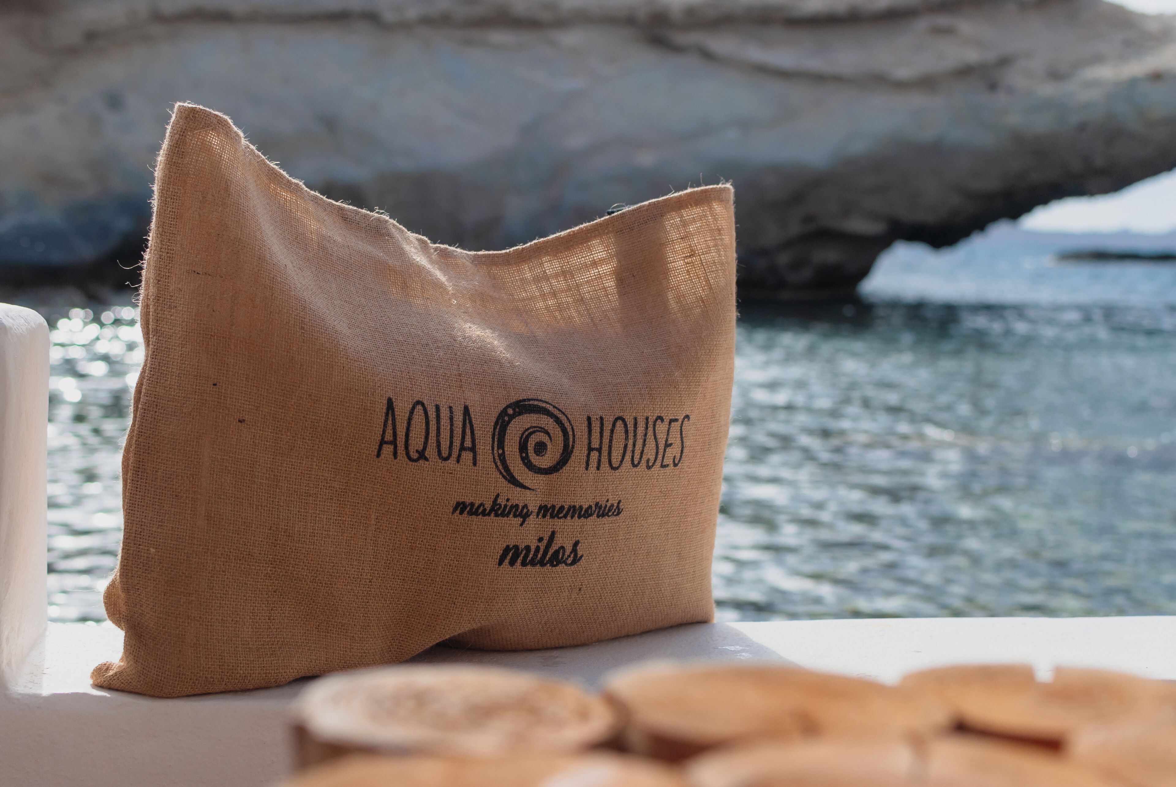 Una bolsa de arpillera con el texto "AQUA HOUSES making memories milos" sobre una mesa cerca del agua.