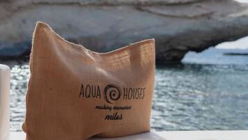 Una bolsa de arpillera con el texto "AQUA HOUSES making memories milos" sobre una mesa cerca del agua.