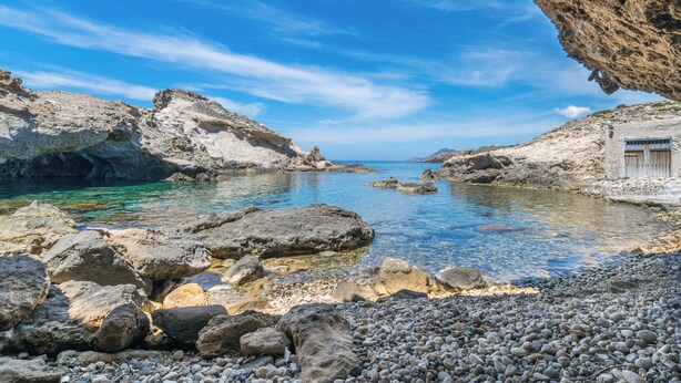 A rocky coastal landscape with clear water, a small building, and a large rock formation. - Aqua Houses (Milos)