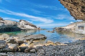 Paisaje costero rocoso con aguas claras, un edificio pequeño y una gran formación rocosa.