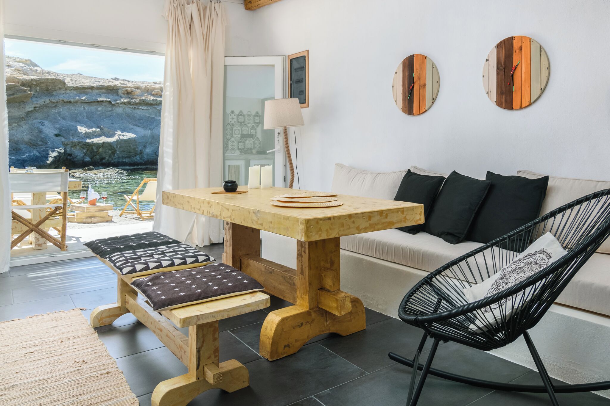 A cozy room with a wooden table, bench, and black chair, a sofa with cushions, and a view of the sea through a glass door.