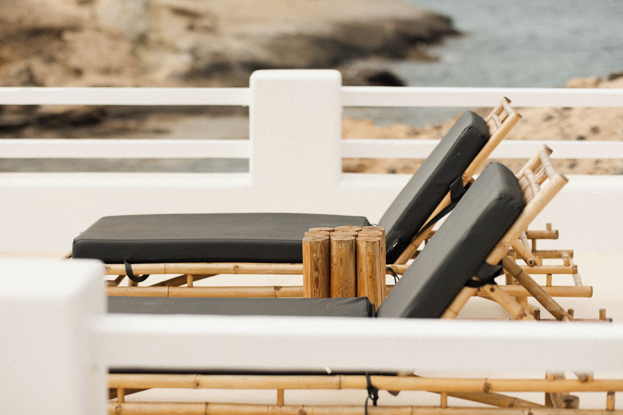 Una silla de playa con cojín negro, estructura de madera y una barandilla blanca al fondo.
