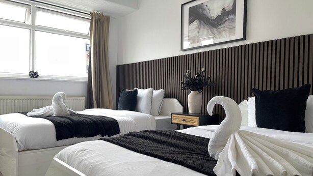 A hotel room with two beds, a large window with curtains, a wall-mounted painting, a bedside table with a vase, and a radiator. - Stylish Woodhouse Home Near Leeds – Sleeps Ten with Parking Included (Leeds)