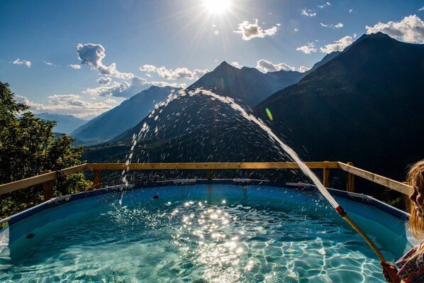 A swimming pool with a wooden deck, surrounded by mountains and a clear sky.