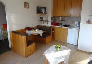 A compact kitchen with wooden cabinets, a microwave, a stove, and a sink. There is a small table with a vase and a chair with a cushion.
