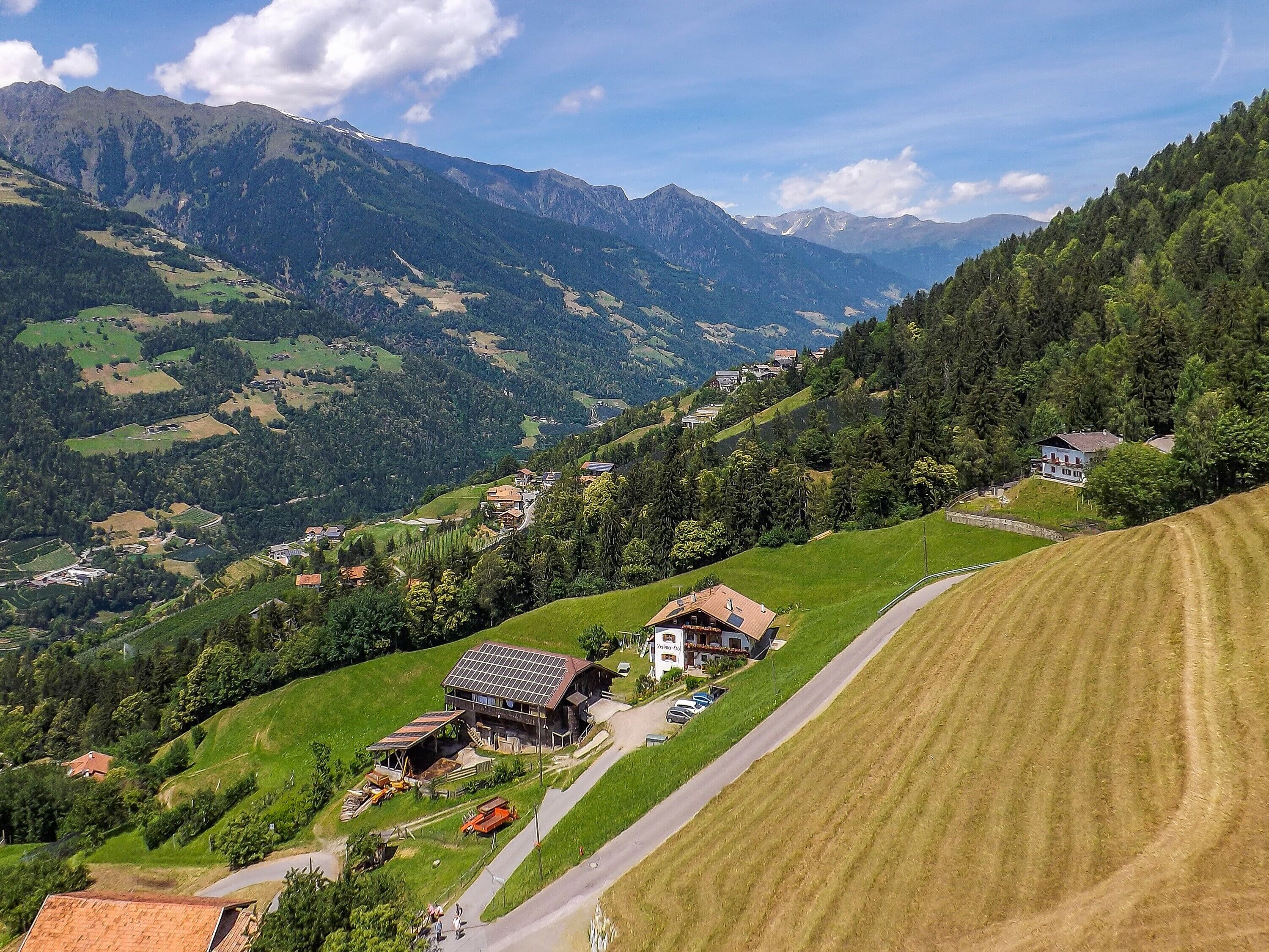 Paisagem montanhosa rural com um pequeno edifício que parece ser uma hospedaria ou pousada, cercada por campos e árvores.